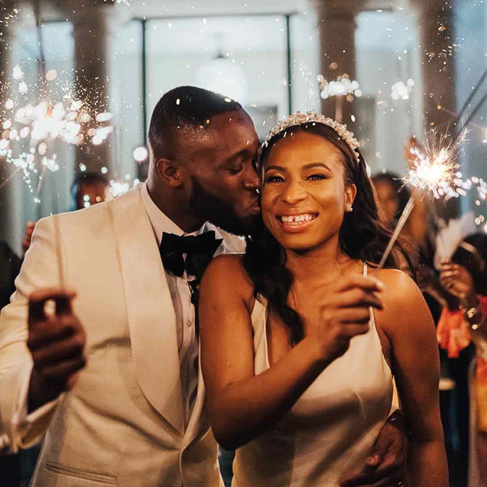 Bride in sleeveless satin wedding dress and pearl headband standing with groom in white tuxedo and black bow tie doing wedding sparkler exit 