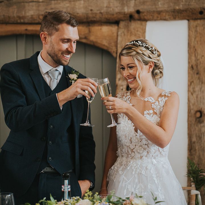 Bride with blonde braided hairstyle with lace wedding dress with groom in navy suit cheers their glasses of champagne together 