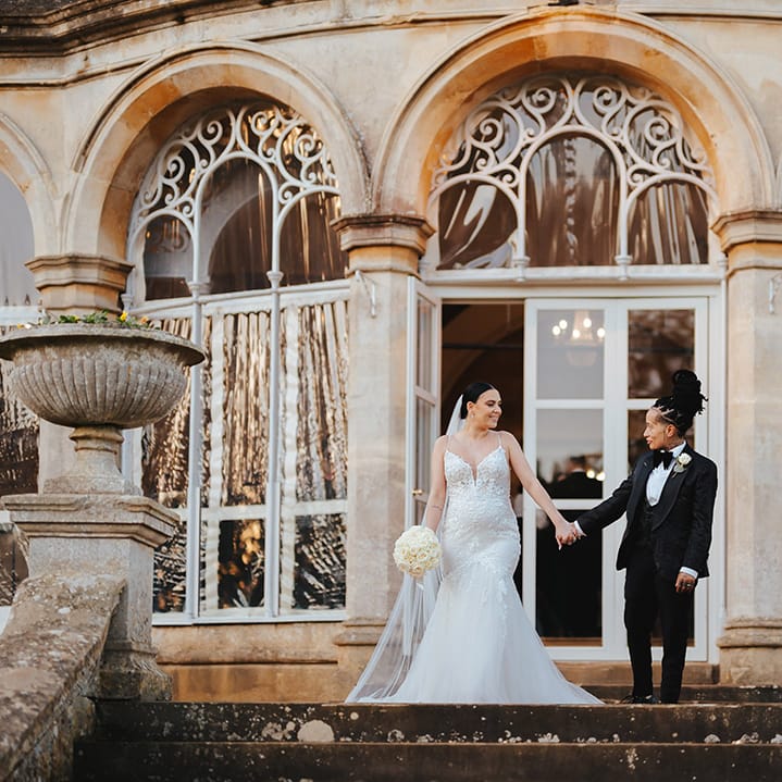 Bride in black snakeskin print tuxedo with black bowtie and white rose boutonniere holding hands with bride in lace sleeveless v neck Enzoani wedding dress with tulle finish holding white garden rose bouquet outside Grittleton House wedding venue 