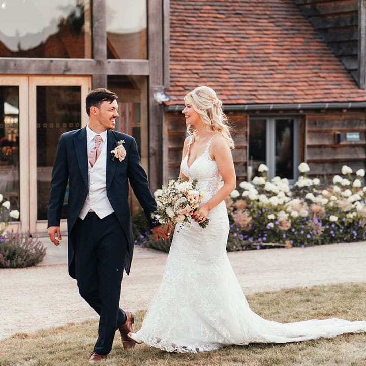Groom in navy morning suit with light grey waistcoat and pink tie walking along with the bride in a lace wedding dress at rustic barn wedding venue 