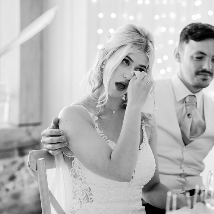 The bride wipes her eyes as she gets emotional listening to the wedding speeches with the groom putting his arm around the bride 