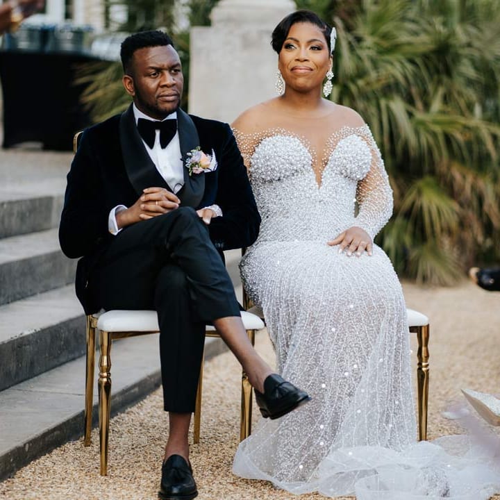 Groom in velvet midnight blue double breasted groom’s blazer, velvet bowtie and peach garden rose boutonniere sitting with bride in illusion v neck sheer off shoulder long sleeve pearl embellished wedding gown at Syon Park vow renewal