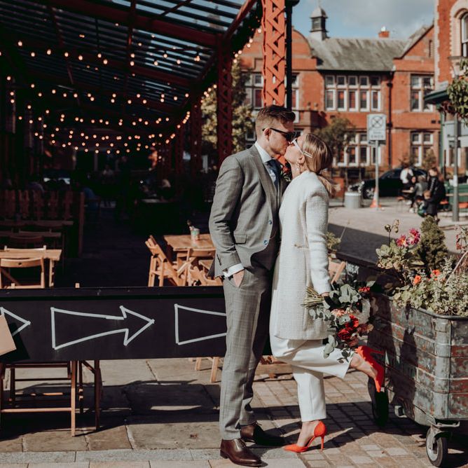 Bride in Massimo Dutti jumpsuit and Ivory coat and groom in grey check suit kissing 