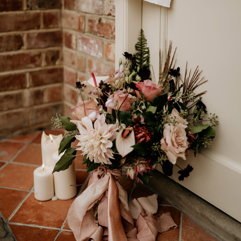 pink flower bouquet with anthuriums, roses and dahlias tied with ribbon leaning up against the glasshouse door with an elopement in progress sign and pillar candles 