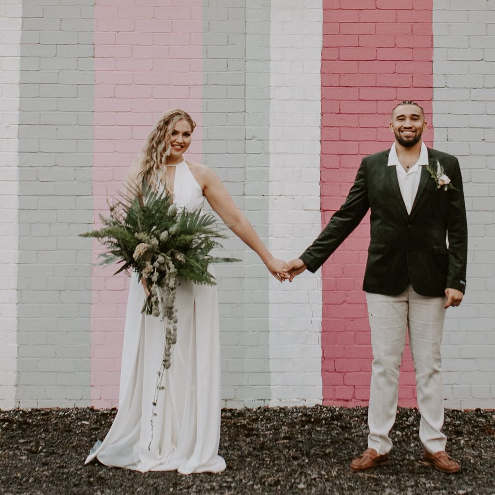 Bride and groom holding hands in front of a pink striped wall, the bride is wearing a wide legged jumpsuit and the groom is wearing a green velvet blazer