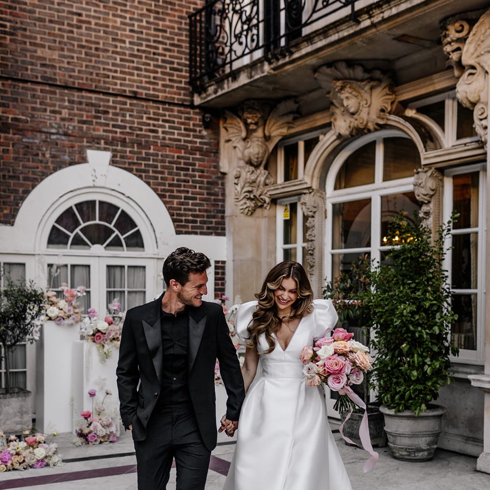 Stylish bride and groom at luxury Mayfair wedding with bride in a Love In Lace Bridal gown with puff sleeves and front split and groom in all black suit and shirt 