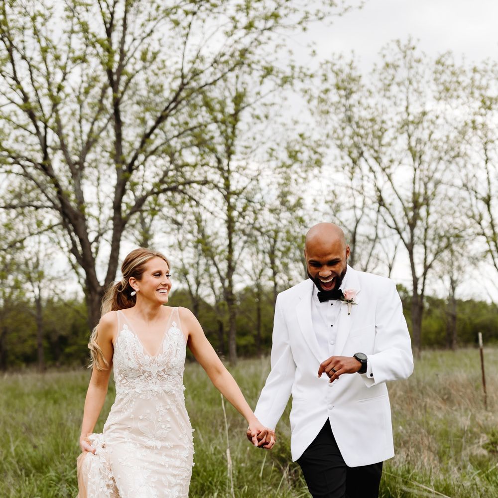 Bride in white embroidered Made With Love wedding dress walks through field holding hands with groom in white tuxedo jacket as they both laugh after backyard wedding 
