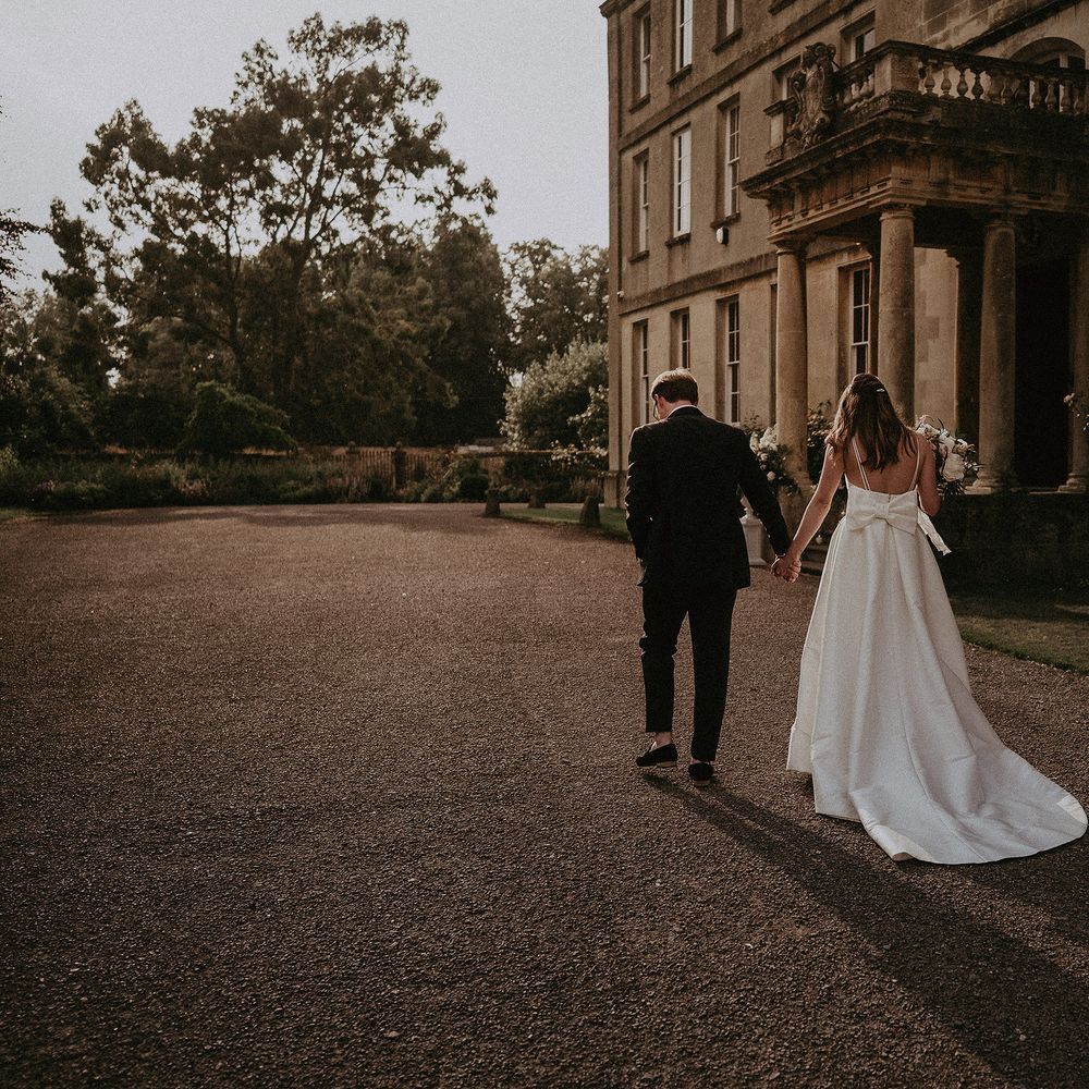 Bride & groom walk hand in hand as bride wears Jesus Peiro dress with bow to the back