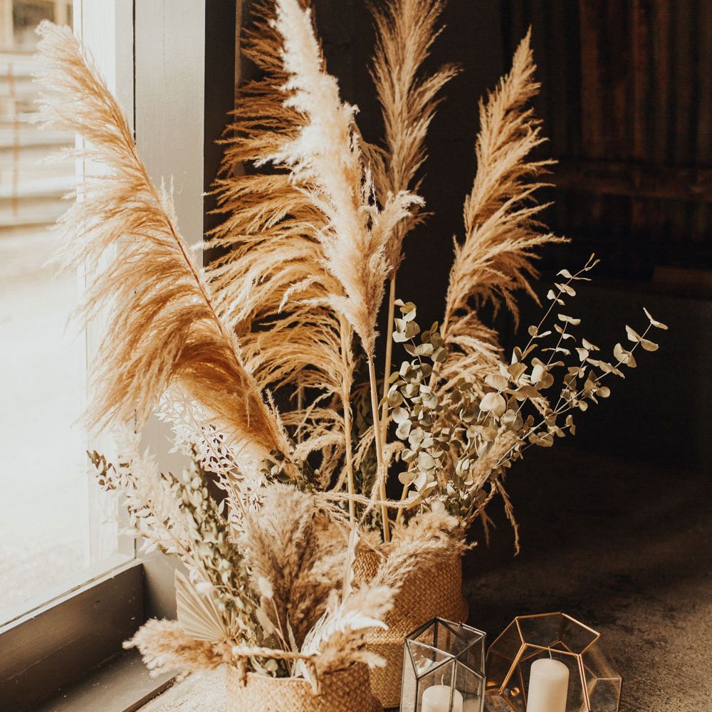 Large pampas grass stems and dried leaves in woven baskets on concrete floor next to metal and glass candle holders with pillar candles for wedding at Anran Devon