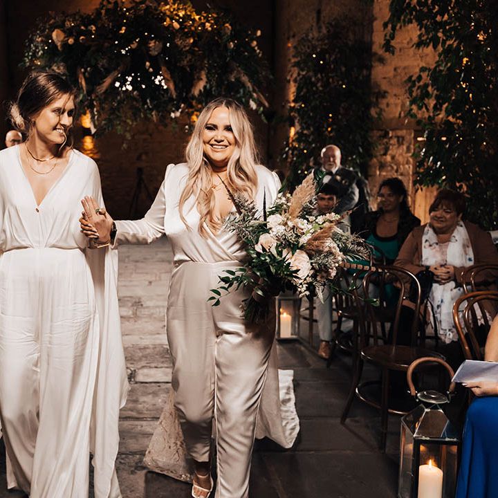 Brides walk down the aisle with one another as blonde bride holds floral bouquet and wedding guests look on whilst smiling