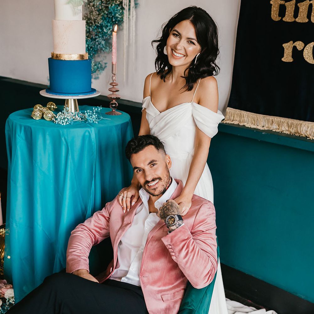 Groom in black trousers, white shirt and pink velvet jacket sitting on a teal velvet chair with his bride in a cold shoulder wedding dress standing behind him 