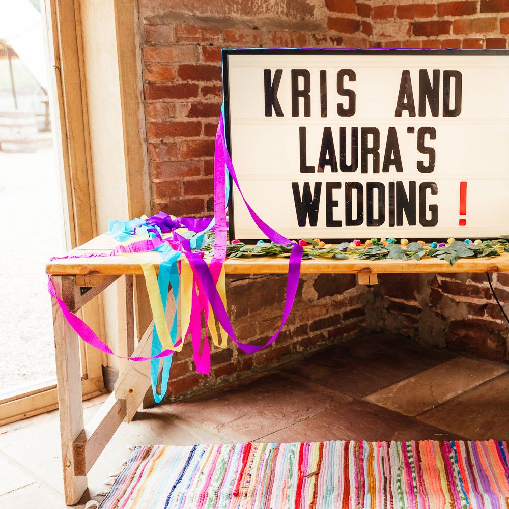 Lightbox wedding sign reading 'Kris and Laura's Wedding!' with streamers and colourful rug for barn wedding 