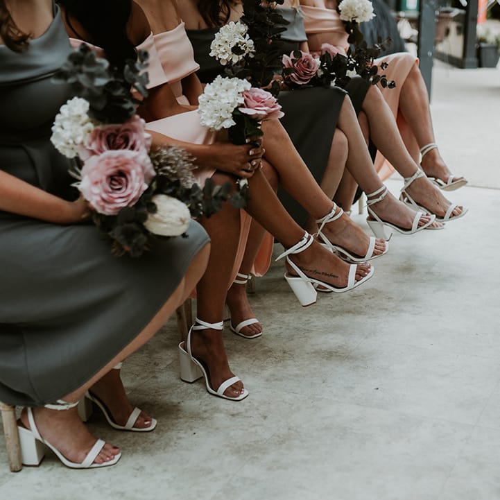 Bridesmaids wear tea length dresses whilst holding pastel bouquets and wearing white strappy block heeled sandals 
