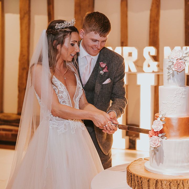 Bride and groom cut into four tier wedding cake with roses and rose gold section in front of light up sign 