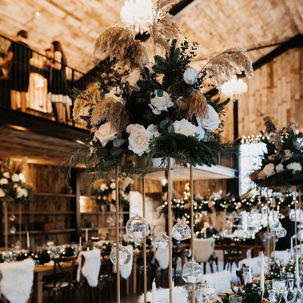 Tall white rose and dried flower centrepieces with hanging glass candles and pinecone runner with white candles table setting