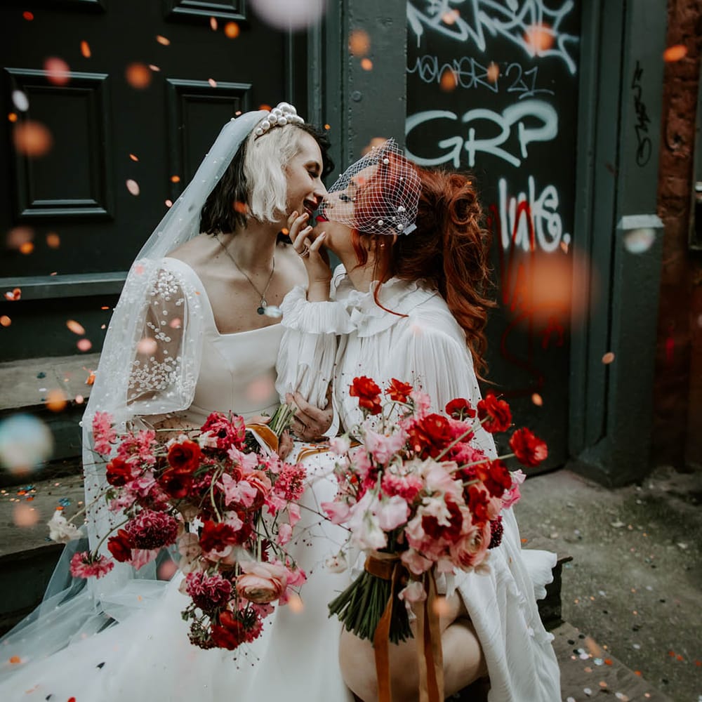 Confetti moment with two brides kissing in a short wedding dress long veil and long sleeve wedding dress and birdcage veil with pearls 