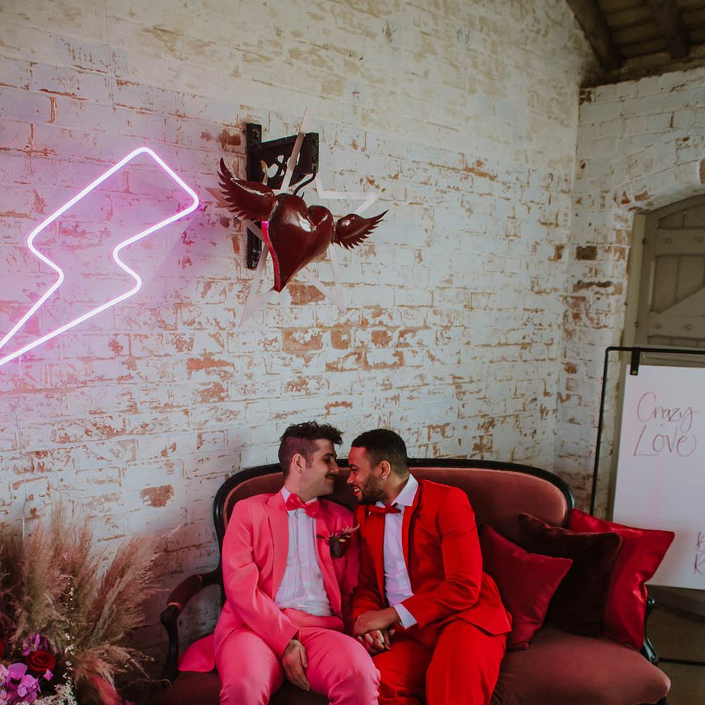 two grooms in pink and red suits sitting on an ornate velvet couch under a pink neon lightning bolt sign and heart with wings 