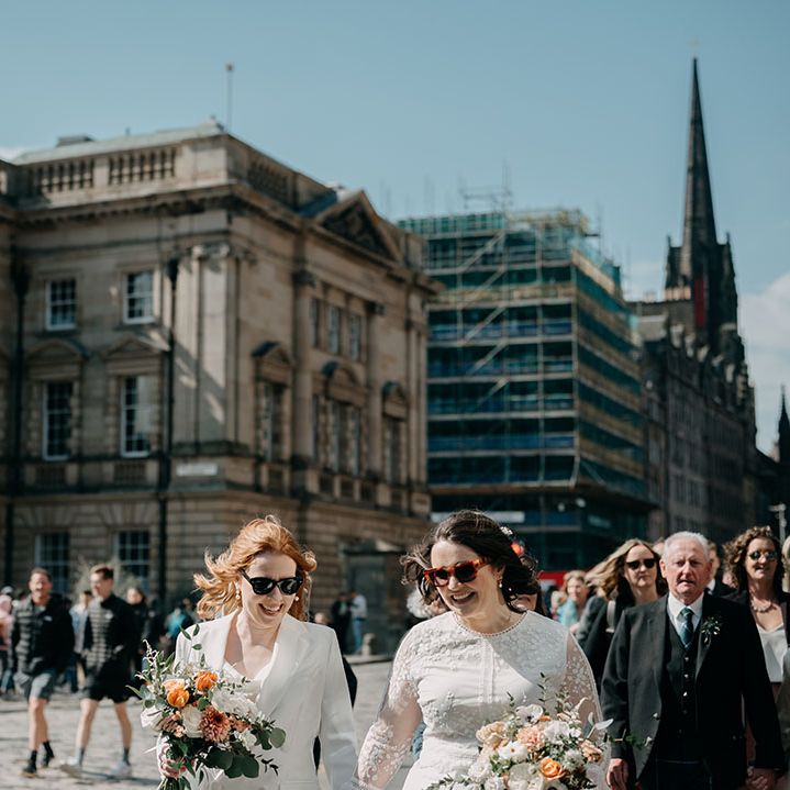 Brides hold hands and walk with one another along the Royal Mile in Edinburgh as family member plays bagpipes 