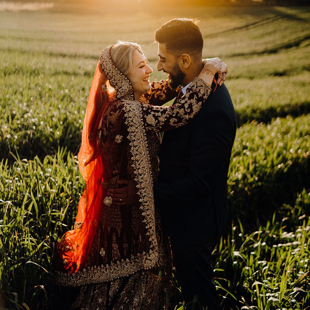 Bride wears traditional saree with deep red colour and gold embellishment whilst stood with her groom during golden hour in field