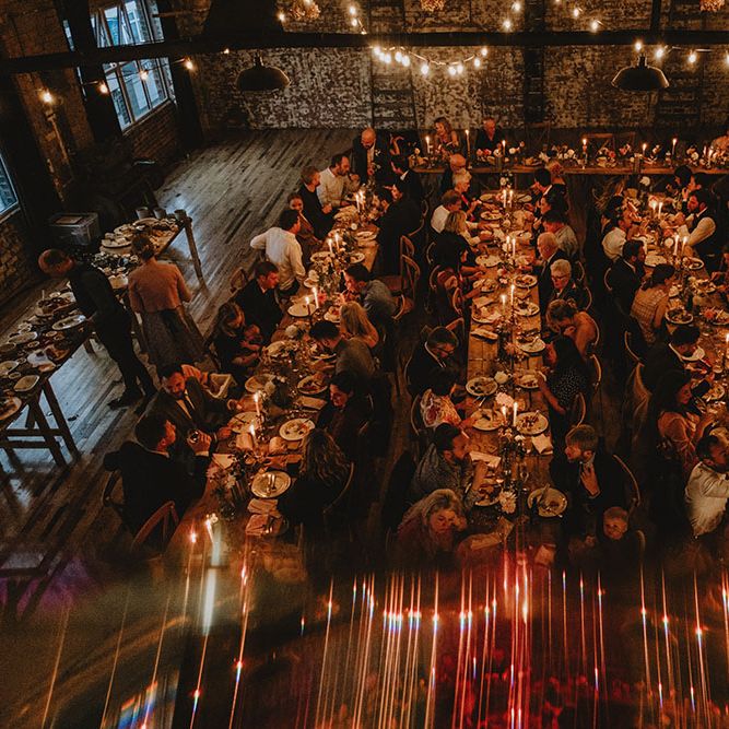 Guests sit at banquet styled wooden tables at Scale Liverpool with industrial feel and exposed brick wall finished with hanging fairy lights