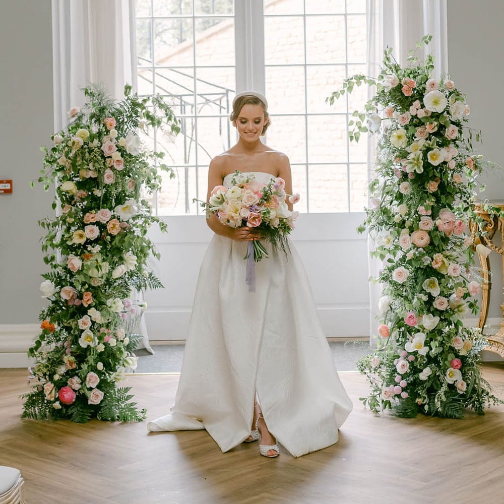 Bride stands inbetween pastel flower columns by Grace Florals holding a matching bouquet in a strapless wedding dress with a headband 
