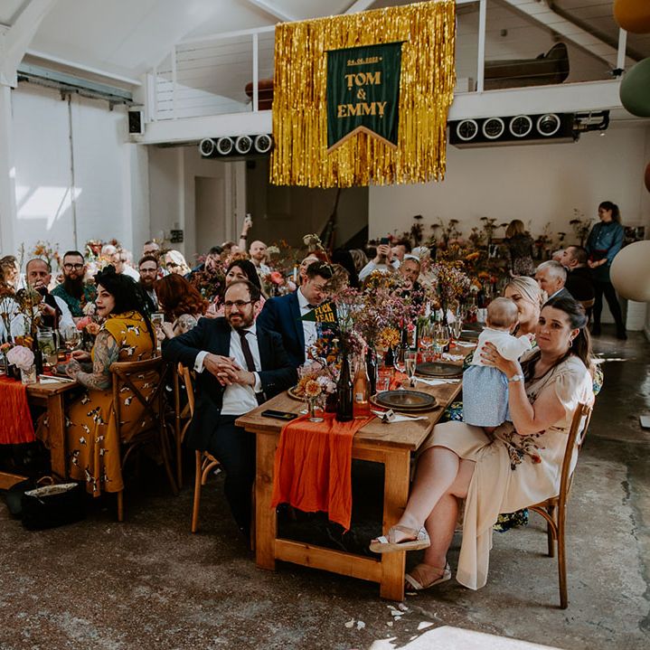 Gold streamers hang from ceiling with velvet banner as wedding guests sit at wooden banquet tables 