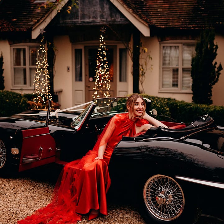 Bride in satin red wedding dress with cape and 3d appliqué roses sitting in a classic convertible wedding car