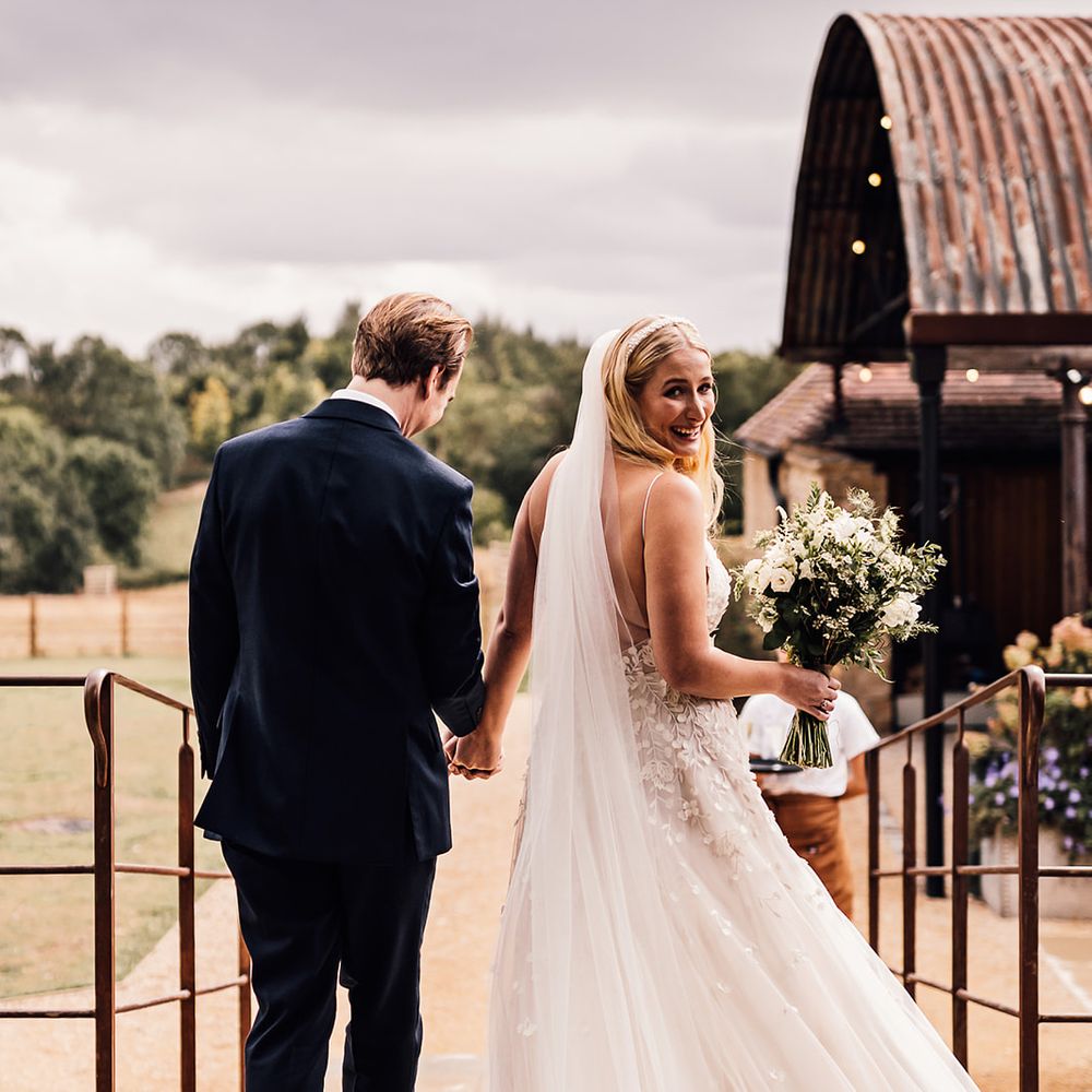 Groom in a dark blue suit walks holding hands with the bride in an appliqué wedding dress 