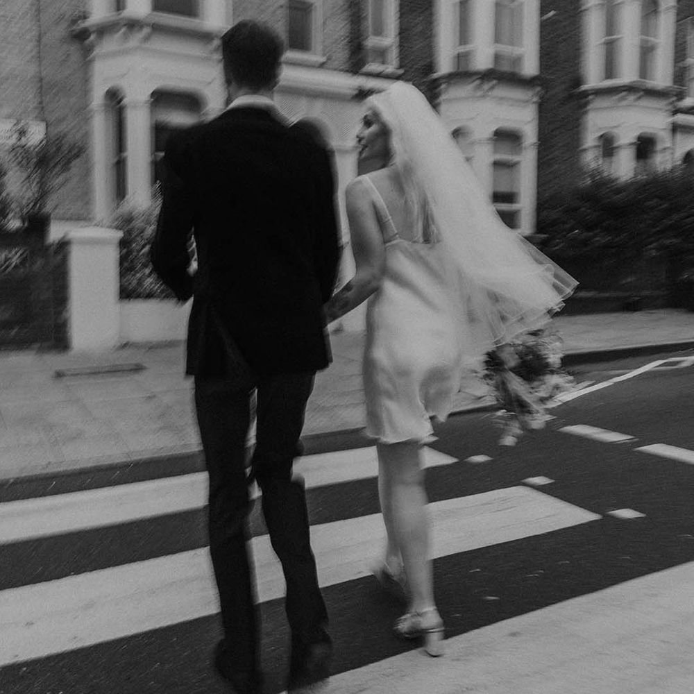 Bride in short satin dress with feather trimmed neckline and groom in dark tux with bowtie holding hands and walking