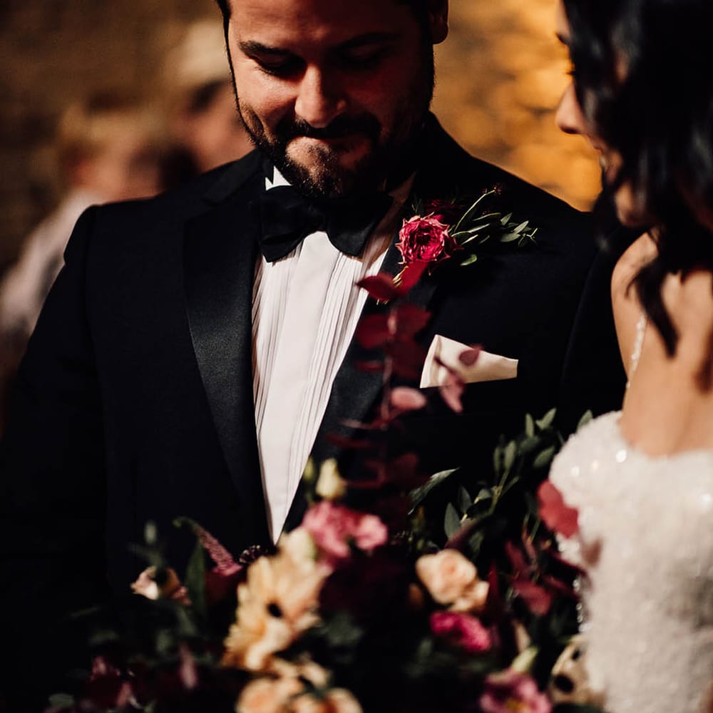 Smiling groom in black tie with a deep pink buttonhole smiling as the bride joins him at the altar 