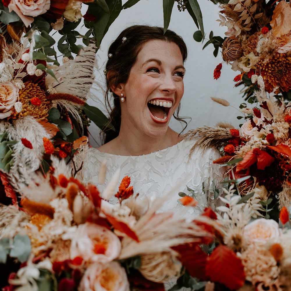 Bride in long sleeve lace vintage wedding dress with pearl earrings and pearl hair clips standing in large autumnal floral arch with roses, peonies, foliage and pampas grass