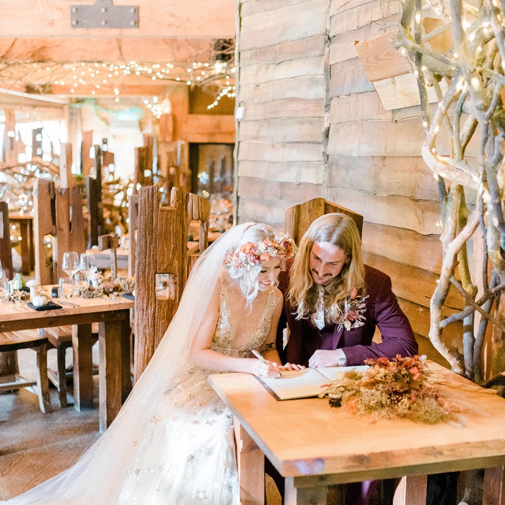 Bride in flower crown and veil sits beside her groom in maroon suit to sign marriage certificate 