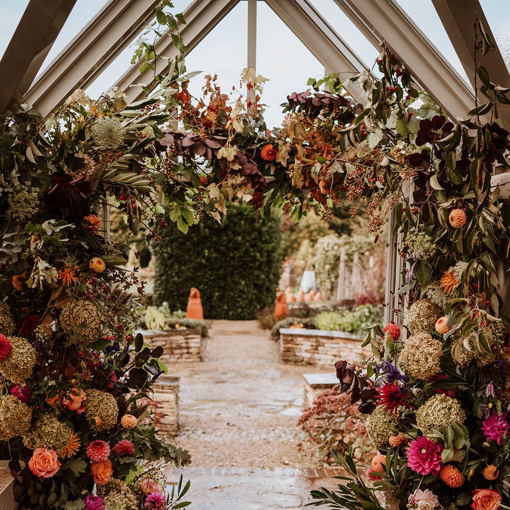 Large luxe floral arch in glasshouse wedding venue with white and pink carnations, garden roses, red strawflower, eucalyptus, baby’s breath and dried flowers 