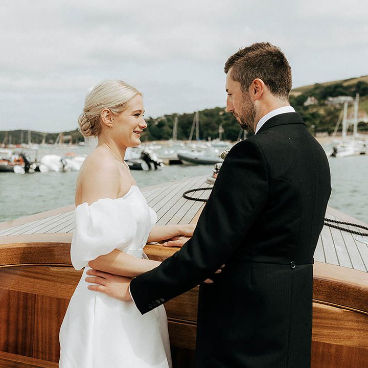 Bride in puffed sleeve Jesus Peiro wedding dress stands beside her groom during boat road for Salcombe wedding 