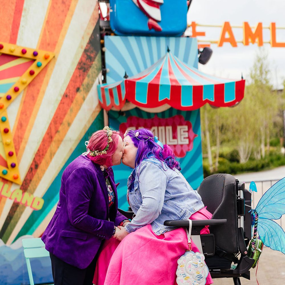 Bride in personalised denim jacket and bright pink wedding dress with blue tights in a wheelchair with sparkly blue fairy wings kissing bride in purple velvet blazer and black suit trousers with purple bowtie, pink patterned pocket square and colourful patterned shirt