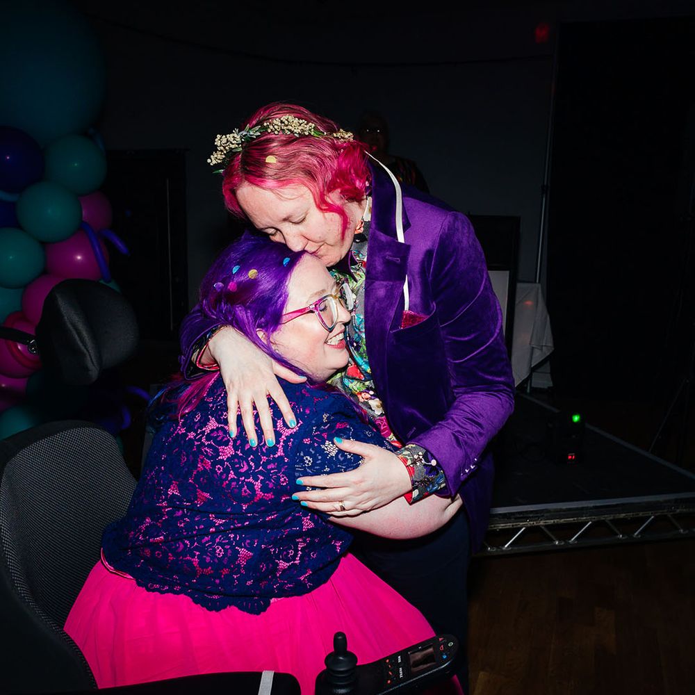 Bride in purple velvet blazer and black suit trousers with purple bowtie, pink patterned pocket square and colourful patterned shirt embracing bride wearing personalised denim jacket, pink wedding dress and blue tights at wedding party 