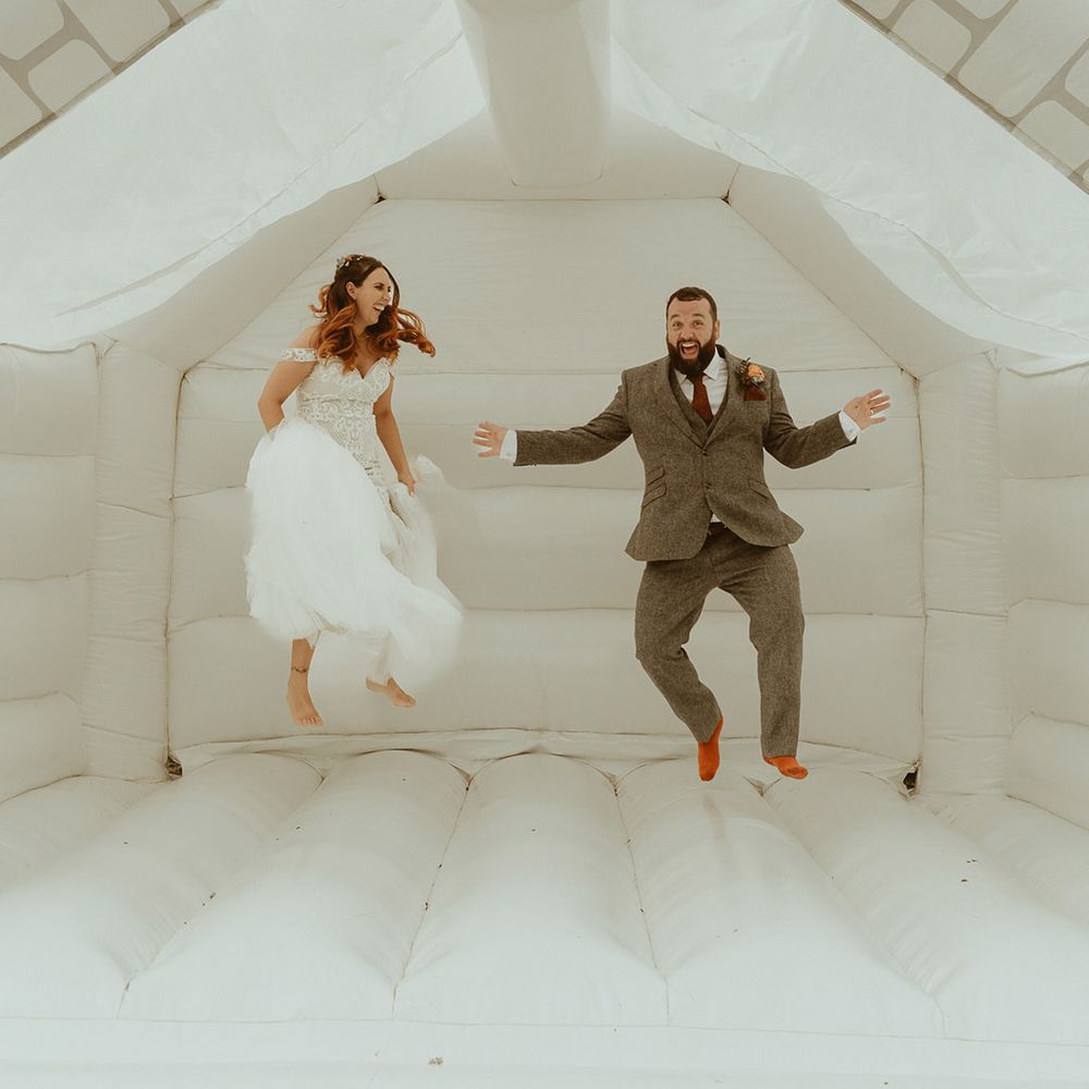 Groom in three piece tweed suit in orange tie with the bride in a fitted lace boho wedding dress jumping on white bouncy castle for bouncy castle wedding 