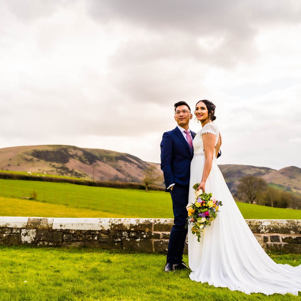 Bride in open back lace wedding dress embraces her groom outdoors surrounded by lush rolling hills 