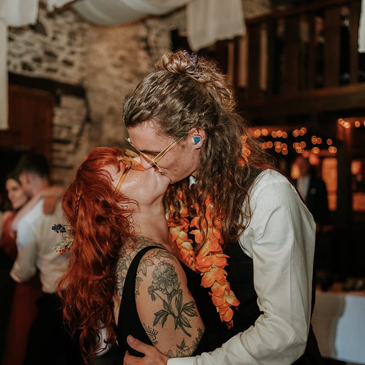 Groom in checkered waistcoat, white shirt and orange lei kissing bride in black wedding dress and orange wedding sunglasses 