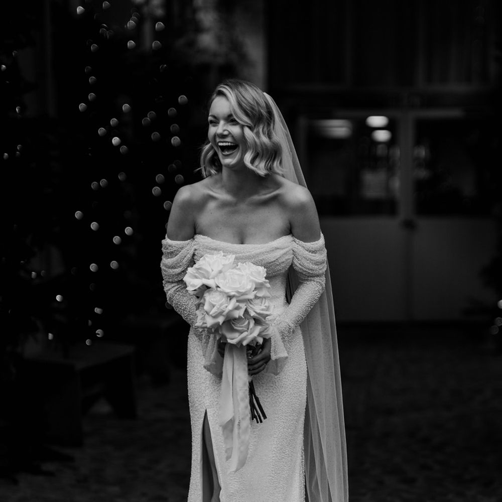 Bride laughing in Emma Beaumont  of the shoulder sparkly wedding dress holding white rose bouquet