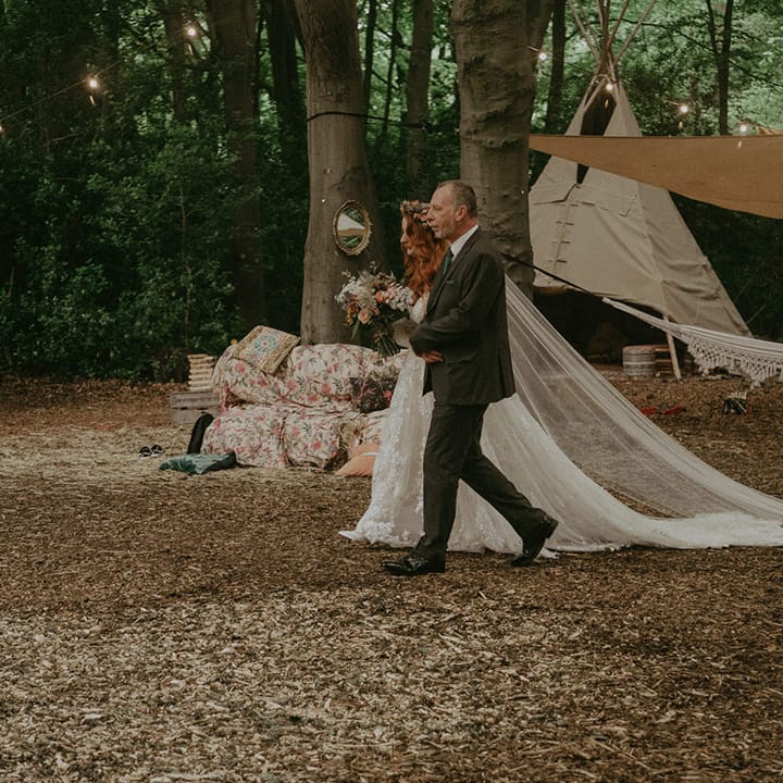Father of the bride in a tweed suit walks the bride in lace wedding dress down the aisle at outdoor wedding 
