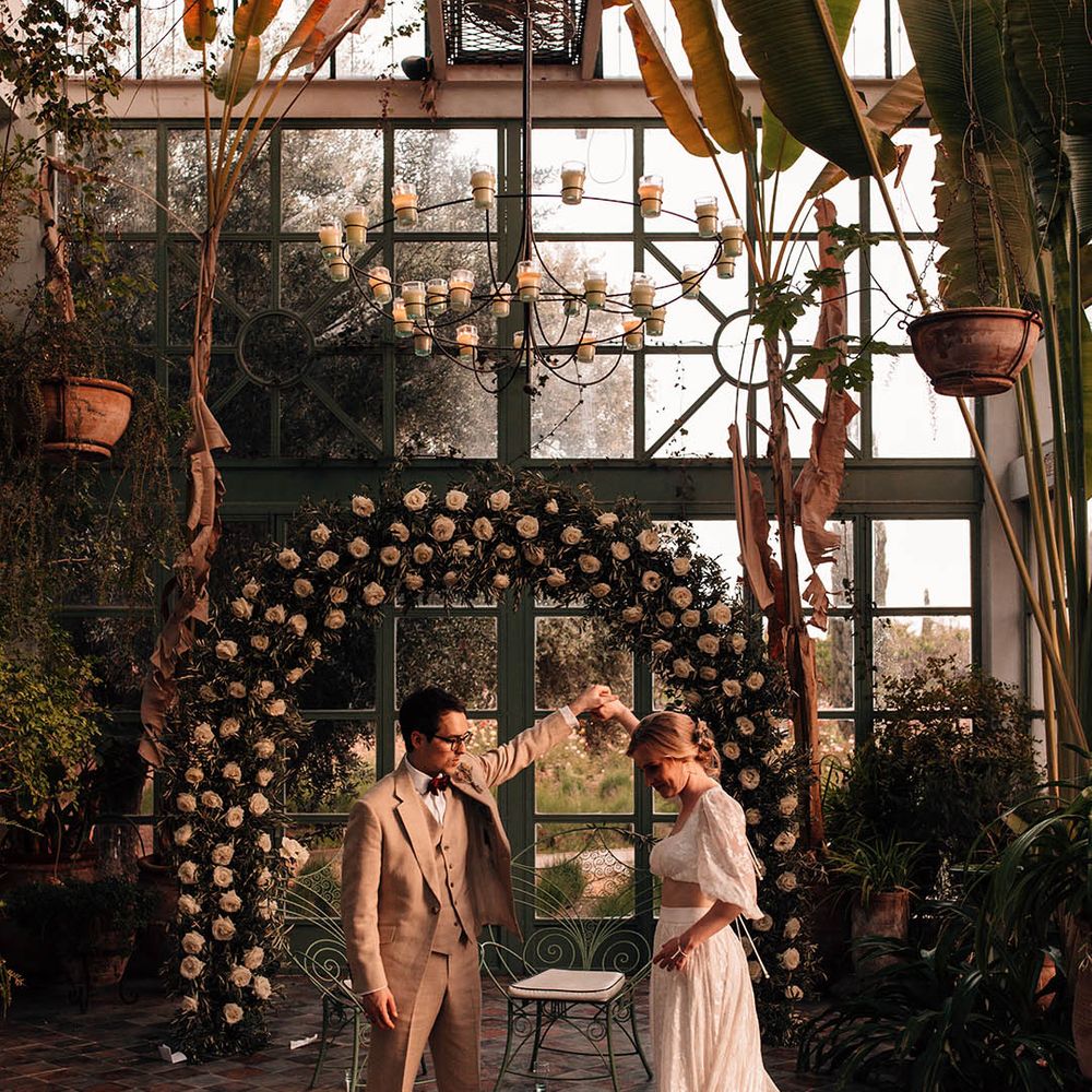 Groom in cream suit spinning the bride round in her modern two piece in front of the white rose arch decoration during golden hour 