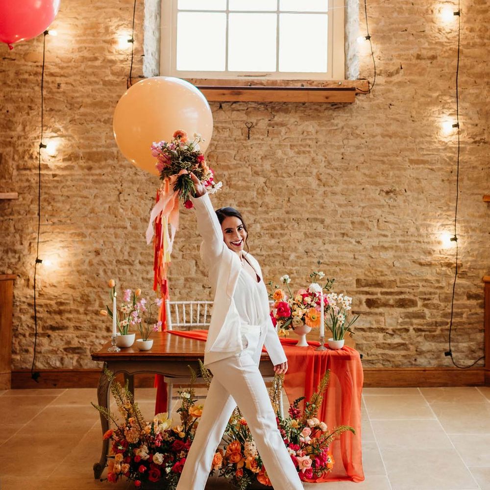Bride in white bridal suit with closed toe bright green wedding heels holding large wedding balloon decoration in the reception room of Merriscourt wedding venue with reception room with metallic pink, white, orange and yellow balloons, wedding fairy lights decorations and wooden table with orange table runner and mixed seasonal flower arrangements 