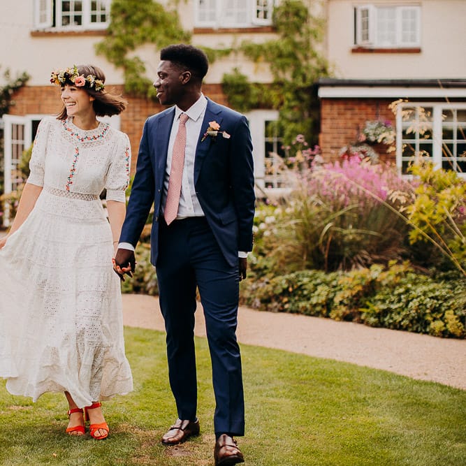 Groom in a navy suit and pink tie holding hands with his bride in a white Broderie Anglaise wedding dress as they arrive for their outdoor wedding reception