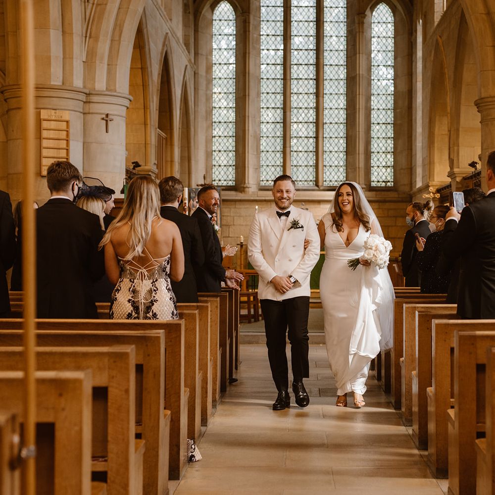 Smiling groom in white tuxedo jacket walks back down the aisle with smiling bride in white Made With Love wedding dress and white chapel length veil