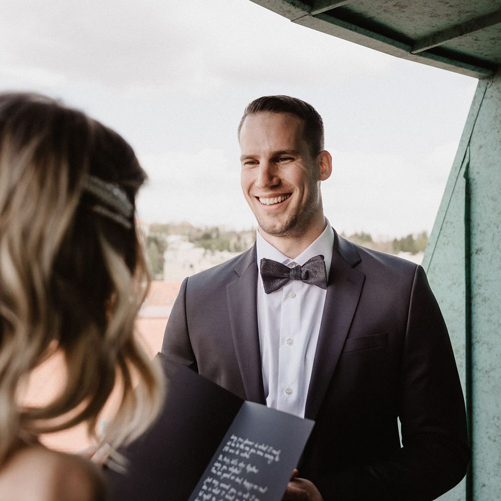 The groom wearing a black suit with white shirt and bowtie, smiling at the bride as she reads her vows