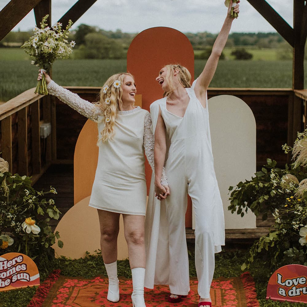 two brides raising their wildflower bouquets in the air standing at the retro inspired altar 