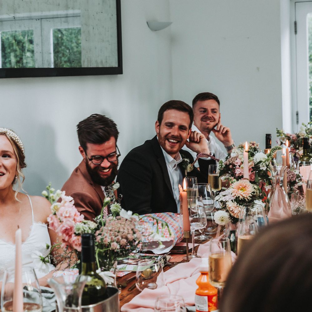 Wedding guests gather around table for wedding reception meal as they laugh and listen to speeches 
