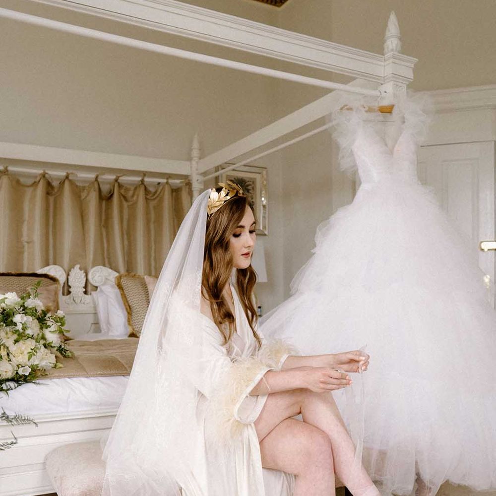 Bride on the wedding morning in a getting ready robe and lace veil putting on her embellished wedding shoes with her tulle wedding dress handing on the four poster bed 