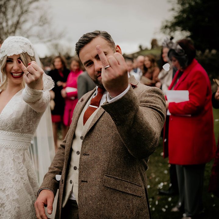 Bride in a lace wedding dress and Juliet cap veil and groom in a wool suit holding up their ring fingers 